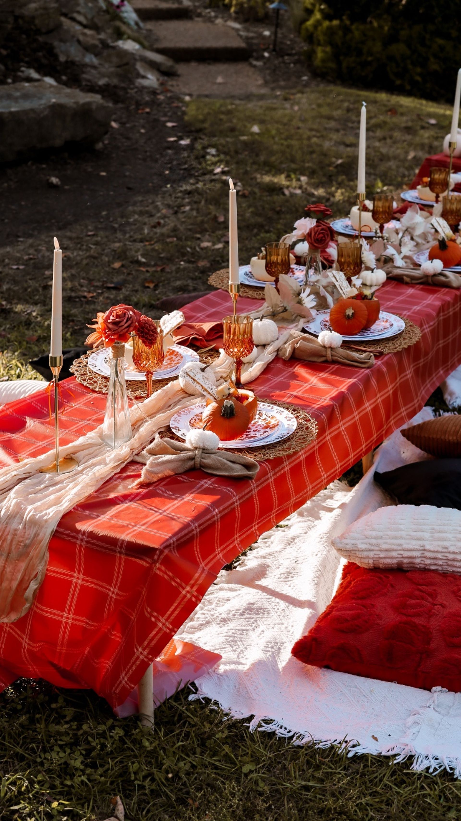 Cozy outdoor autumn tablescape on a red plaid tablecloth in a backyard — pumpkin place settings with woven chargers, amber goblets, tall white taper candles, floral accents and plush floor cushions.