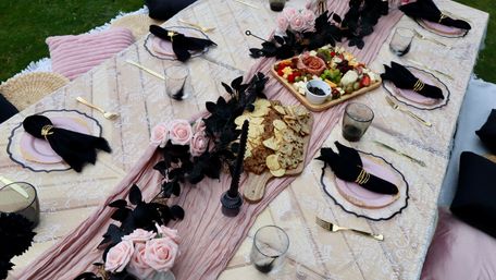 Chic outdoor garden picnic table with pink lace runner, black napkins with gold rings, gold flatware, wooden charcuterie and cracker boards, and blush rose accents