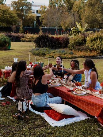 Friends toasting at a cozy autumn garden picnic — low table with orange plaid tablecloth, candles, pumpkins, plates and pillows on the grass.