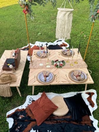 Boho outdoor picnic on green lawn with low wooden tables, layered place settings, gold flatware, taper candles, floral centerpiece, macramé hanging, vintage record player, cowhide rugs and textured pillows.