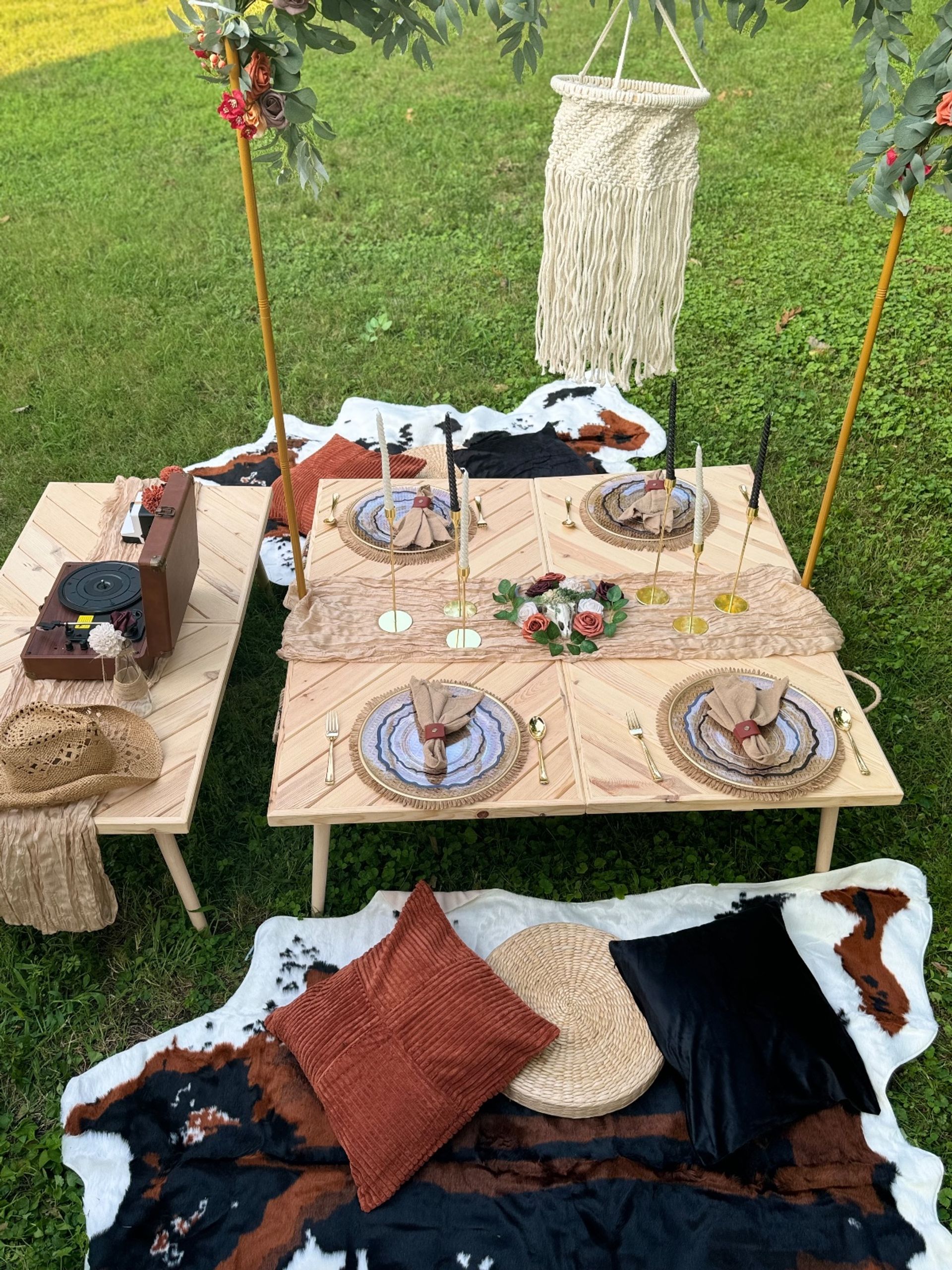 Boho outdoor picnic on green lawn with low wooden tables, layered place settings, gold flatware, taper candles, floral centerpiece, macramé hanging, vintage record player, cowhide rugs and textured pillows.
