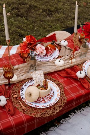 Cozy outdoor autumn picnic spread on red plaid tablecloth with woven placemats, patterned plates, small white pumpkins, floral vases, tall taper candles and a pumpkin bowl of snacks