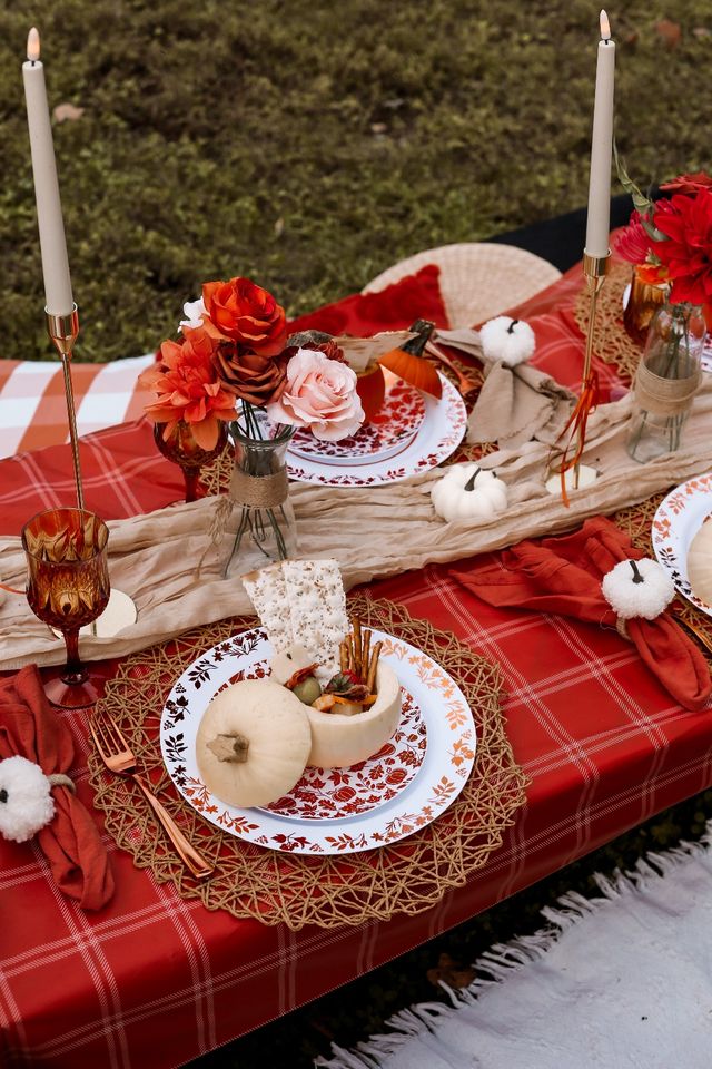 Cozy outdoor autumn picnic spread on red plaid tablecloth with woven placemats, patterned plates, small white pumpkins, floral vases, tall taper candles and a pumpkin bowl of snacks