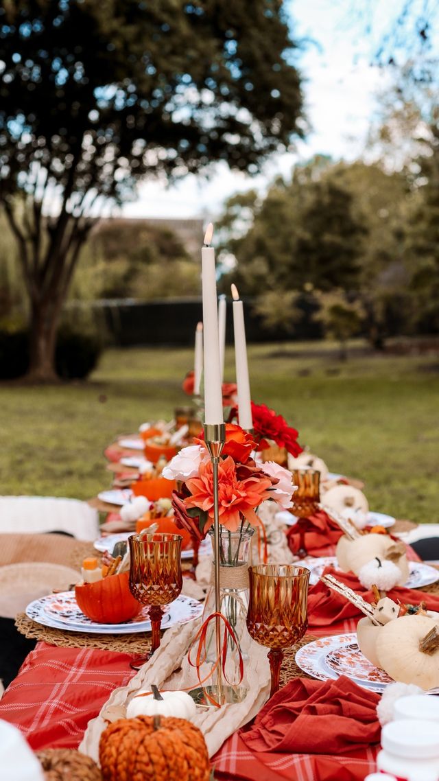 Cozy outdoor fall tablescape with tall white taper candles, amber goblets, orange and white pumpkins, and vibrant orange florals on a red plaid runner for a harvest-style picnic