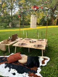 Cozy boho backyard picnic setup with low wooden tables, woven place settings, gold candlesticks, floral garland arch and macramé hanging over cowhide blanket and cushions on green lawn