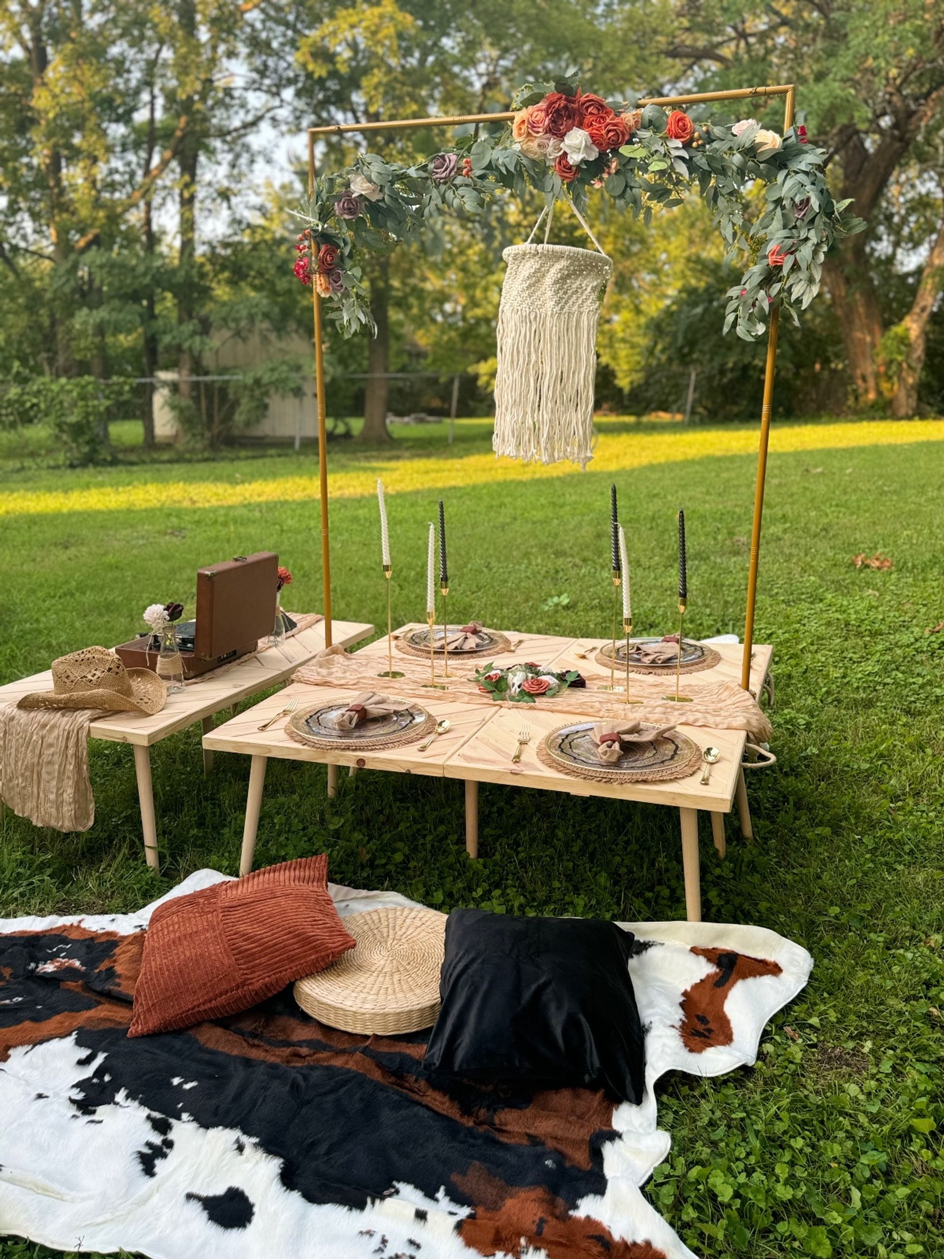 Cozy boho backyard picnic setup with low wooden tables, woven place settings, gold candlesticks, floral garland arch and macramé hanging over cowhide blanket and cushions on green lawn