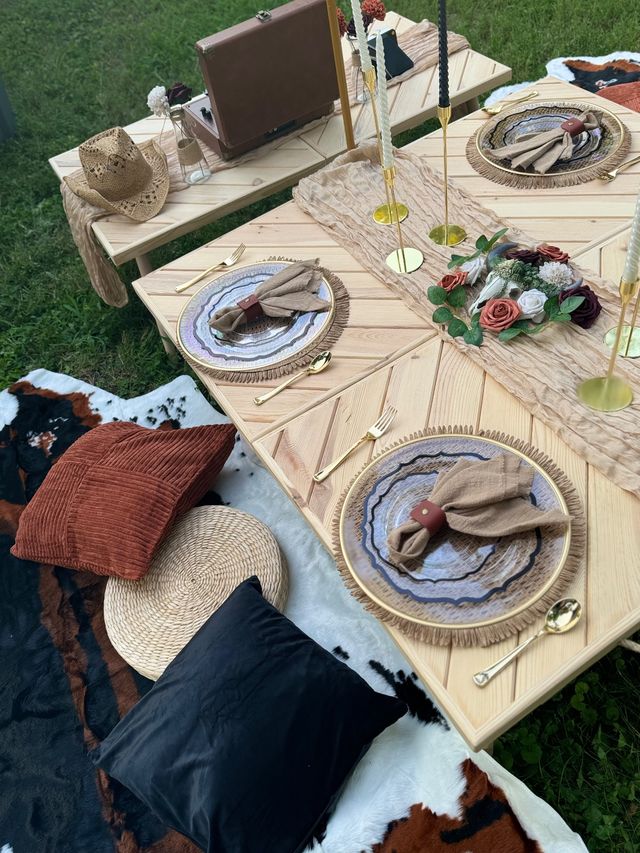 Boho outdoor backyard picnic tablescape on grass with low wooden tables, layered decorative plates and fringed chargers, knotted linen napkins with leather rings, gold flatware, tall brass candle holders, floral-decorated skull centerpiece, straw hat and textured cushions on a cowhide rug.