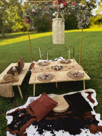 Cozy boho outdoor picnic on a green lawn with low wooden tables set for four, macramé hanging lantern, floral arch, taper candles, woven chargers, cowhide throw and textured cushions
