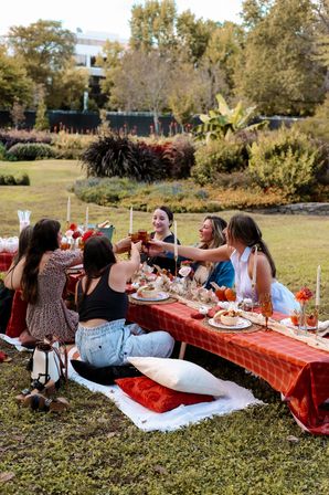 Friends clinking glasses at a cozy outdoor fall picnic in a park — low table with orange plaid tablecloth, candles, pumpkins, seasonal table settings and cushions on the grass with garden trees in the background.
