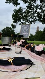 Playful bachelorette picnic in a park: decorated table with blush roses, black napkins and gold rings, glassware and a framed 'R.I.P. Last Name' sign under leafy trees with a distant tower in the background.