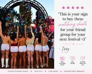 Group of friends at an outdoor music festival seen from behind, wearing matching custom white foldover shorts and colorful tops with arms raised toward a floral-decorated arch and illuminated stage at dusk