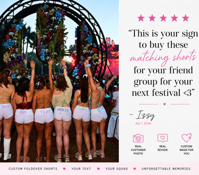 Group of friends at an outdoor music festival seen from behind, wearing matching custom white foldover shorts and colorful tops with arms raised toward a floral-decorated arch and illuminated stage at dusk