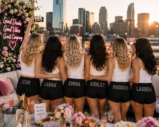 Sunset rooftop skyline celebration: six friends in white tops and matching black personalized shorts clinking pink cocktails and taking a selfie amid floral decor and a neon sign reading Good Drinks Good Vibes Great Company.
