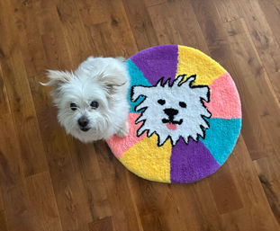 Small fluffy white dog on a hardwood floor next to a round colorful rug featuring a cartoon dog face in teal, purple, yellow, and pink