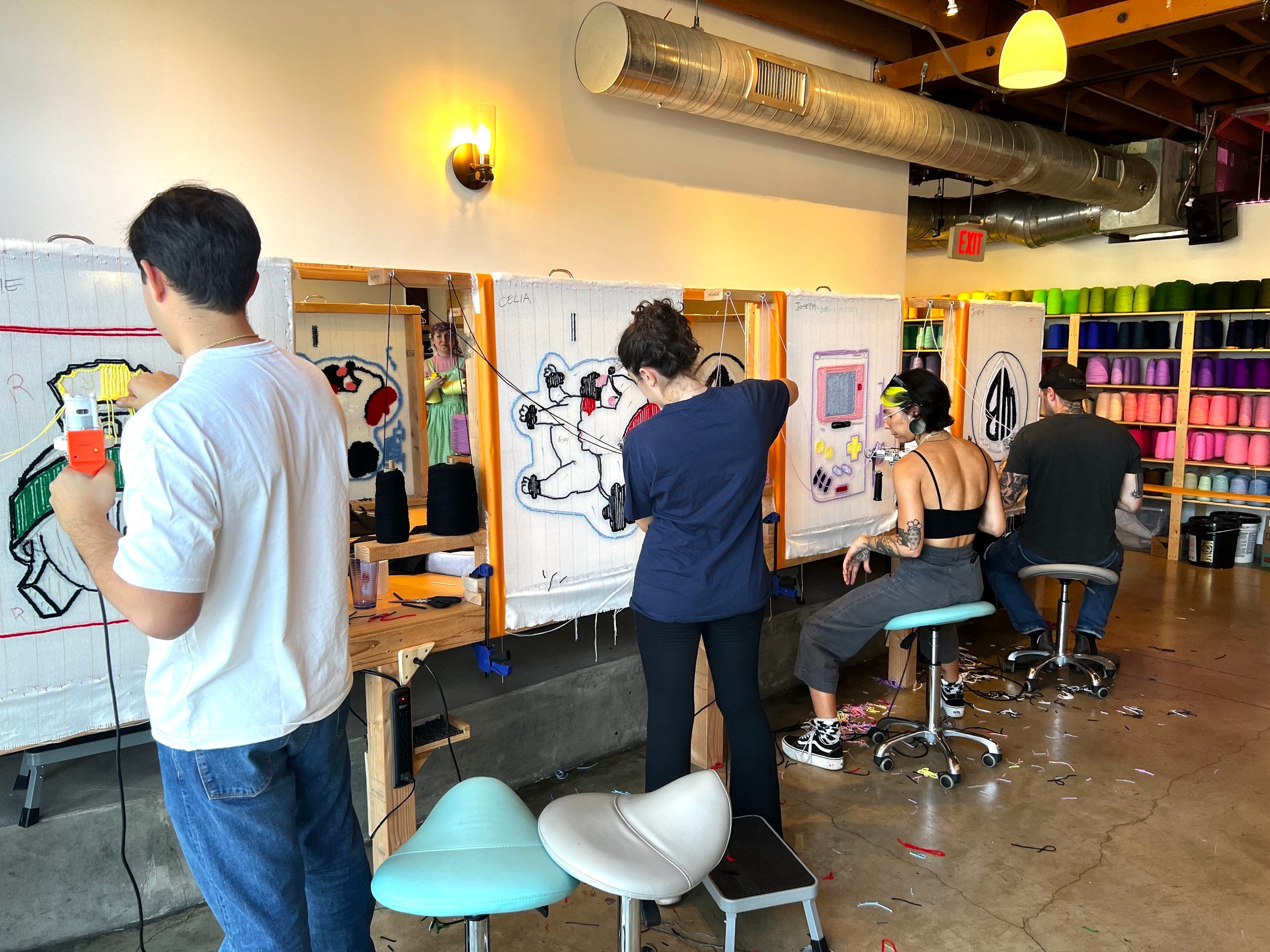 Four people in a bright tufting workshop using tufting guns on framed canvases to create colorful yarn rugs, with shelves of yarn cones, stools and industrial ceiling ducts.