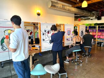 Four people in a bright tufting workshop using tufting guns on framed canvases to create colorful yarn rugs, with shelves of yarn cones, stools and industrial ceiling ducts.