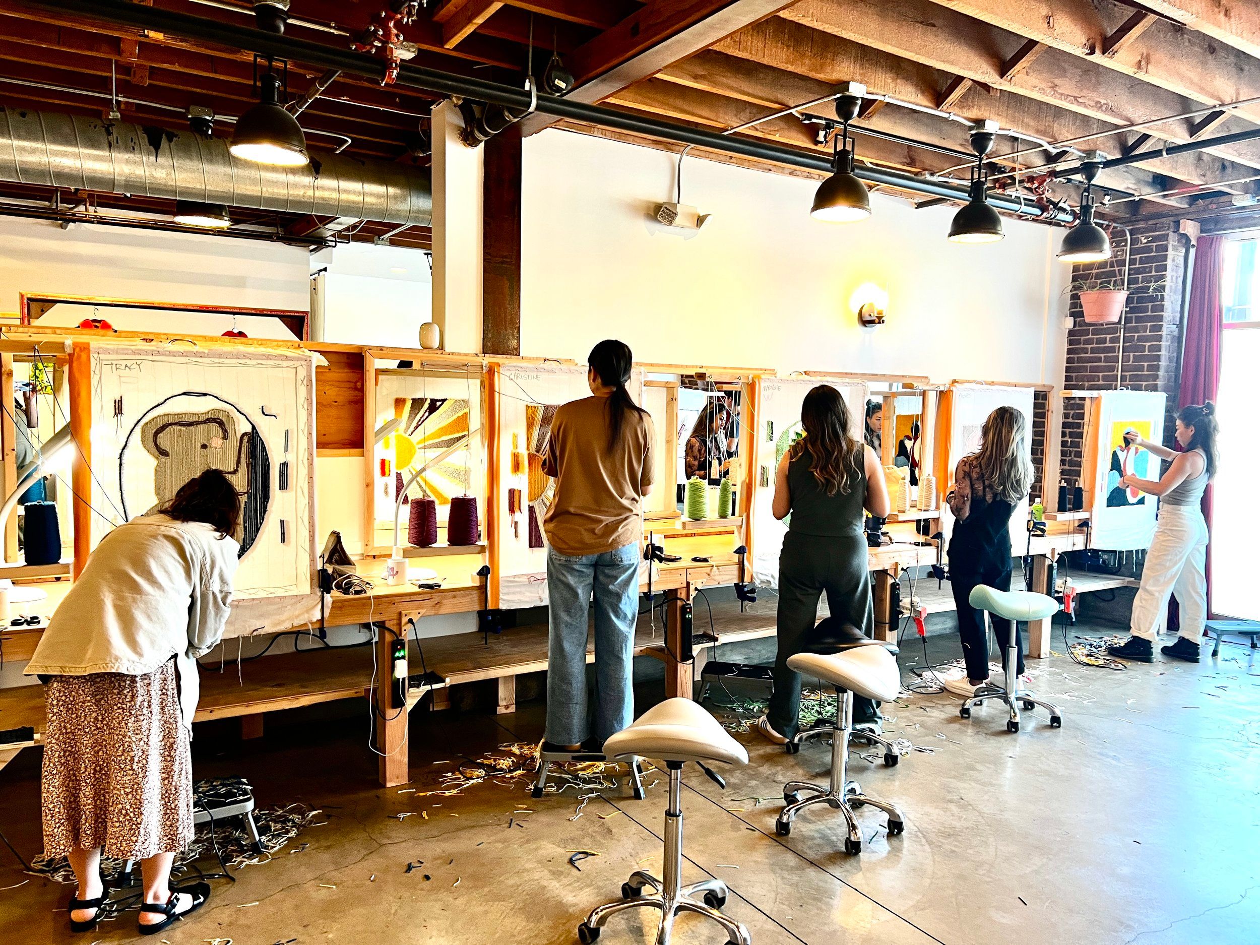 Hands-on group weaving workshop in a sunlit industrial loft: five people at wooden loom stations with colorful yarn cones and mirrors, exposed brick and beams, pendant lights overhead and yarn scraps on the concrete floor.