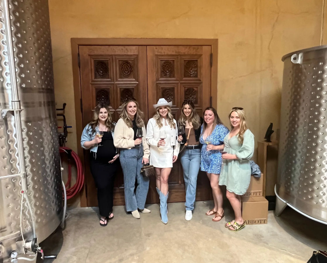 Six women smiling and holding wine glasses in a winery tasting room between stainless-steel fermentation tanks, posed in front of carved wooden doors.