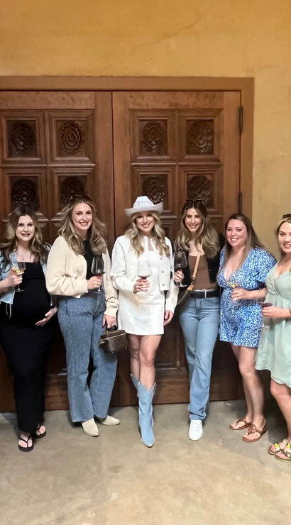 Six women smiling and holding wine glasses in a winery tasting room between stainless-steel fermentation tanks, posed in front of carved wooden doors.