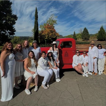 Smiling group of women in white outfits posing by a vintage red pickup truck and hay bales on a sunny countryside patio with tall trees and blue sky