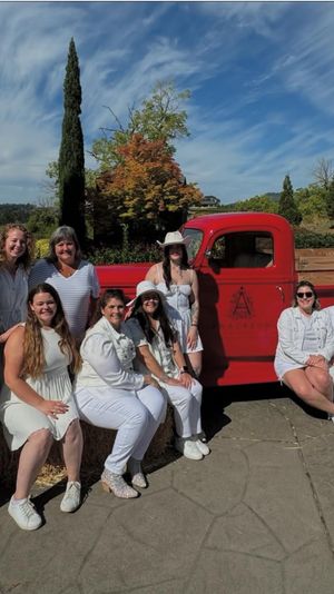 Smiling group of women in white outfits posing by a vintage red pickup truck and hay bales on a sunny countryside patio with tall trees and blue sky