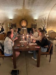 Candlelit wine cellar tasting: a group of friends seated at a long wooden table with wine glasses, barrels, wreath and warm ambient lighting.