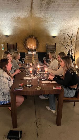Candlelit wine cellar tasting: a group of friends seated at a long wooden table with wine glasses, barrels, wreath and warm ambient lighting.