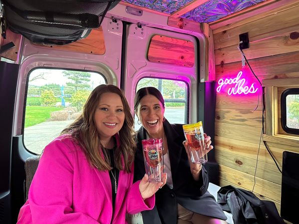 Two smiling women pose inside a cozy wood-paneled camper van lit with pink neon "good vibes" signage, each holding a colorful travel tumbler.