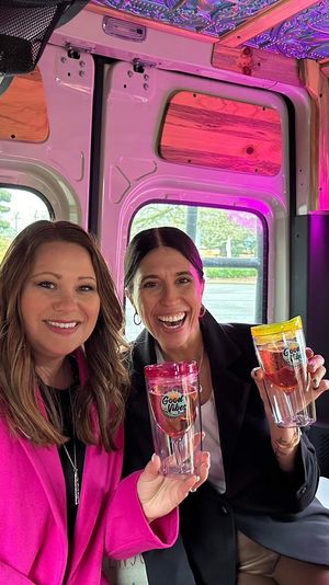 Two smiling women pose inside a cozy wood-paneled camper van lit with pink neon "good vibes" signage, each holding a colorful travel tumbler.