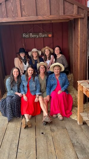 Eight women in sundresses, denim jackets and wide-brim hats smiling while seated on hay bales inside a rustic red barn doorway — casual country-style group portrait.