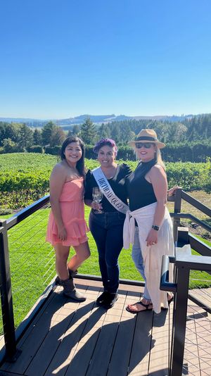 Three women on a sunny winery terrace overlooking grapevines and rolling hills, one wearing a 'Birthday Queen' sash and holding a wine glass during an outdoor celebration.