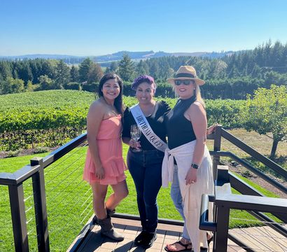 Three women on a sunny winery terrace overlooking grapevines and rolling hills, one wearing a 'Birthday Queen' sash and holding a wine glass during an outdoor celebration.