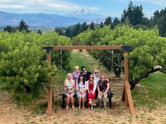 Seven women pose on a wooden swing bench in a lush fruit orchard, framed by rows of leafy trees with forested hills and a distant snow‑capped mountain under a blue sky.