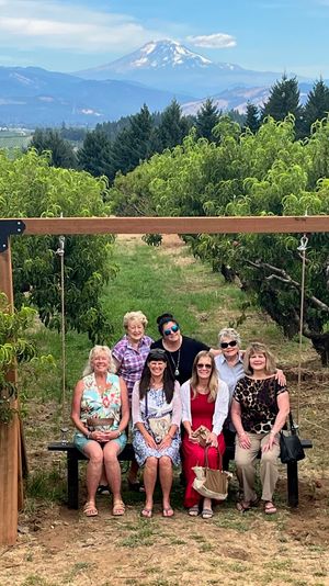 Seven women pose on a wooden swing bench in a lush fruit orchard, framed by rows of leafy trees with forested hills and a distant snow‑capped mountain under a blue sky.