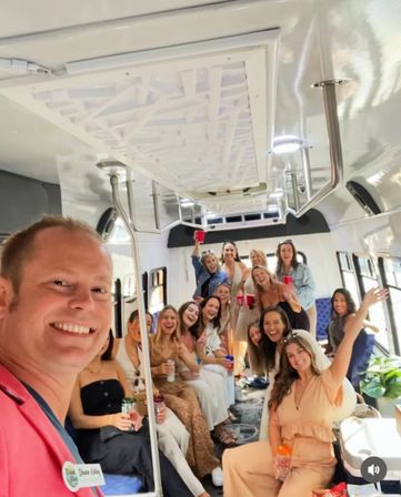 Cheerful group of friends taking a selfie inside a bright white party bus, seated along benches, holding red cups and drinks, stainless-steel poles and overhead lights, festive vibe.
