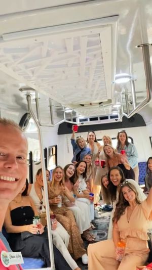 Cheerful group of friends taking a selfie inside a bright white party bus, seated along benches, holding red cups and drinks, stainless-steel poles and overhead lights, festive vibe.