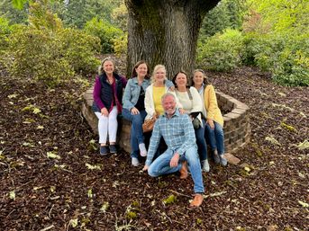 Six smiling adults seated on a curved stone bench around a large oak tree in a leafy park, wearing casual jackets and jeans on a wood-chip ground