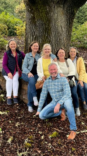 Six smiling adults seated on a curved stone bench around a large oak tree in a leafy park, wearing casual jackets and jeans on a wood-chip ground