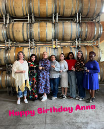Group of eight women holding wine glasses and smiling in a winery cellar, posing in front of stacked oak barrels for a birthday celebration.