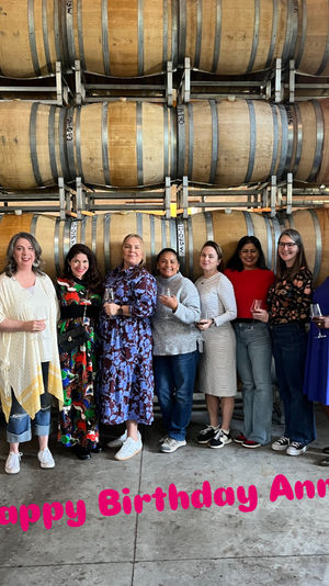 Group of eight women holding wine glasses and smiling in a winery cellar, posing in front of stacked oak barrels for a birthday celebration.