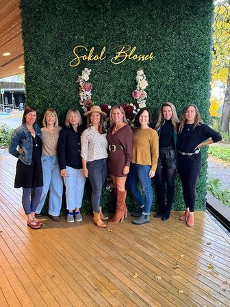Eight smiling women posing on a wooden deck in front of a lush green floral wall with gold cursive signage and pink-and-white flower accents, wearing casual fall outfits, boots and a hat, with autumn trees visible nearby.