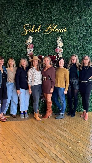 Eight smiling women posing on a wooden deck in front of a lush green floral wall with gold cursive signage and pink-and-white flower accents, wearing casual fall outfits, boots and a hat, with autumn trees visible nearby.