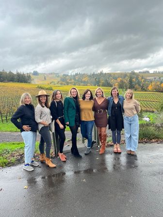 Eight women posing on wet pavement in front of a rolling autumn vineyard with colorful vines and a dramatic overcast sky