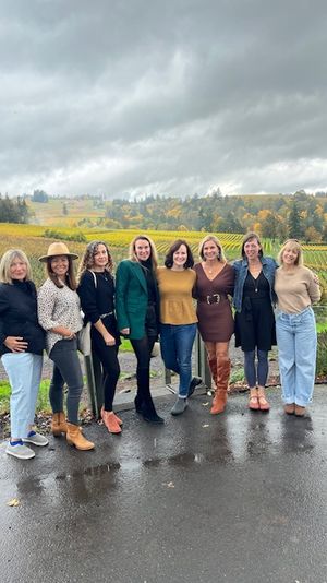Eight women posing on wet pavement in front of a rolling autumn vineyard with colorful vines and a dramatic overcast sky