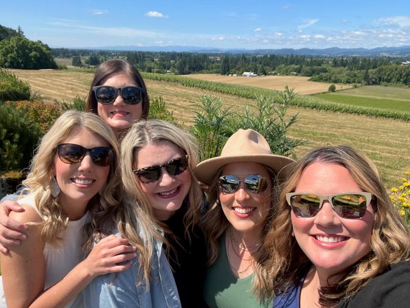 Five friends wearing sunglasses smiling for a selfie on a sunny day with rolling vineyard and farmland under a blue sky.