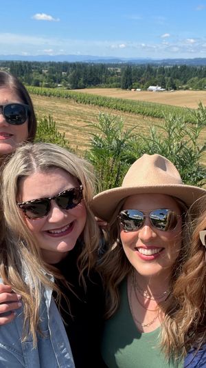 Five friends wearing sunglasses smiling for a selfie on a sunny day with rolling vineyard and farmland under a blue sky.