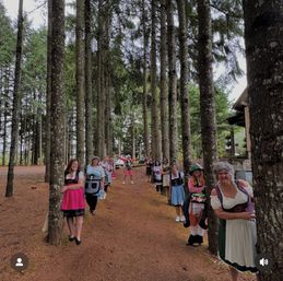 Playful group in dirndl-style dresses lined up peeking from behind tall pine trees along a woodland path, with a cabin and parked car visible in the forest background.