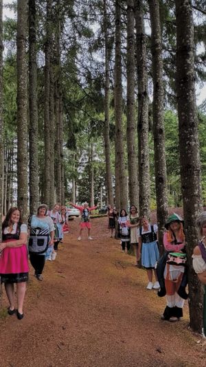 Playful group in dirndl-style dresses lined up peeking from behind tall pine trees along a woodland path, with a cabin and parked car visible in the forest background.