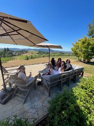 Group of friends sipping wine on a shaded patio overlooking vineyard rows, rolling hills and a bright blue sky