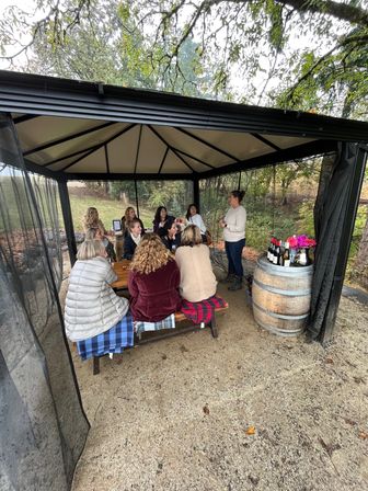 Group of people at an outdoor wine tasting under a screened gazebo on a wooded hillside, seated at picnic tables while a host speaks beside a wine barrel with bottles and flowers — cozy autumn gathering.